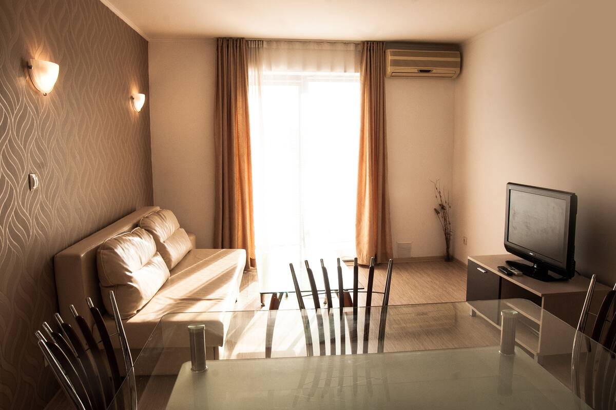 Una habitación de hotel moderna con una mesa de comedor de vidrio, un sofá, un televisor y un ventanal con cortinas.