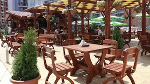 An outdoor café with wooden tables and chairs, potted plants, and a covered seating area. - Rainbow 1 Holiday Complex (Sunny Beach)