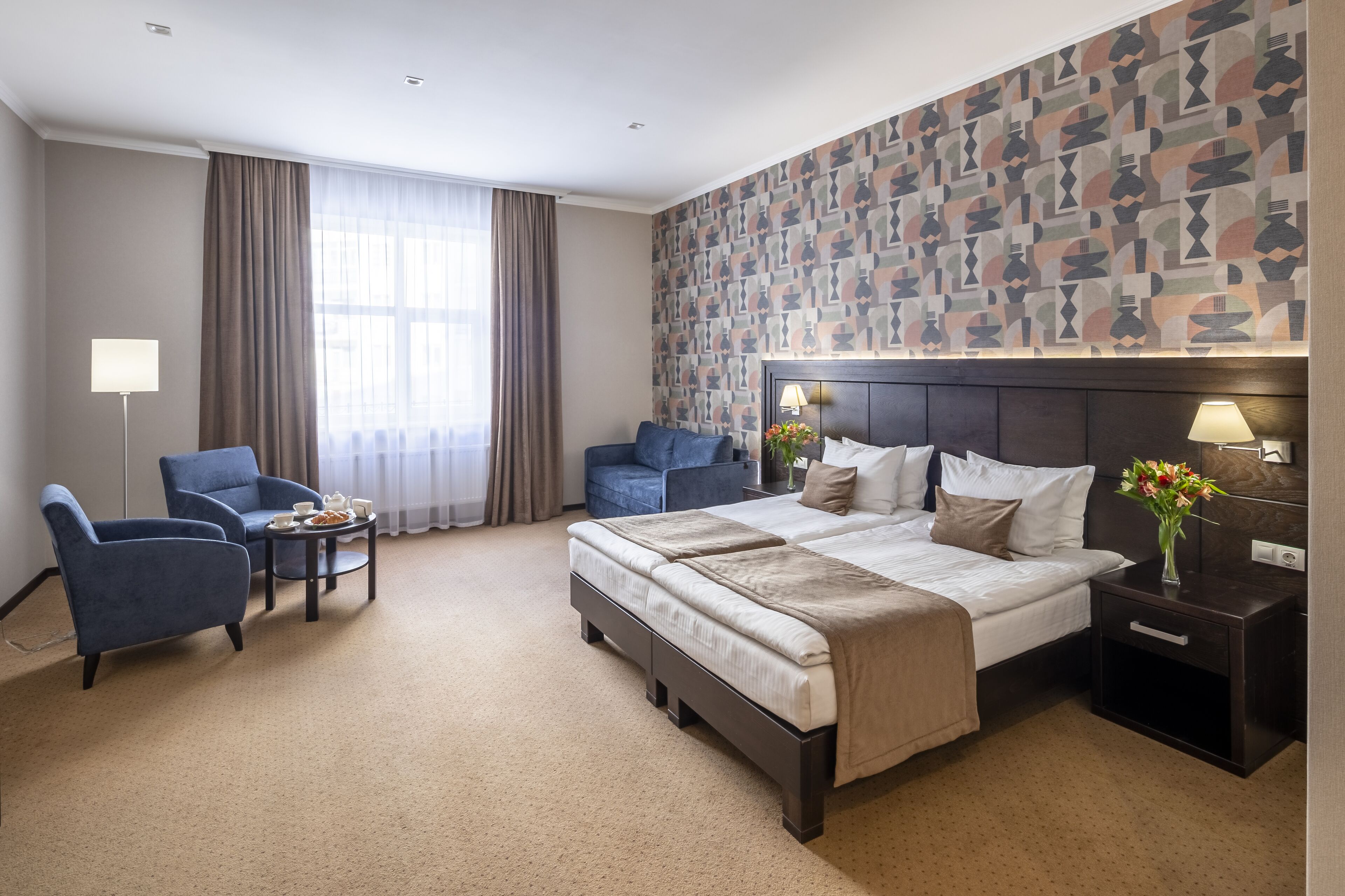 Une chambre d’hôtel avec un grand lit, deux fauteuils, une petite table avec un plateau de nourriture, une table de chevet avec une lampe et un mur à motifs.