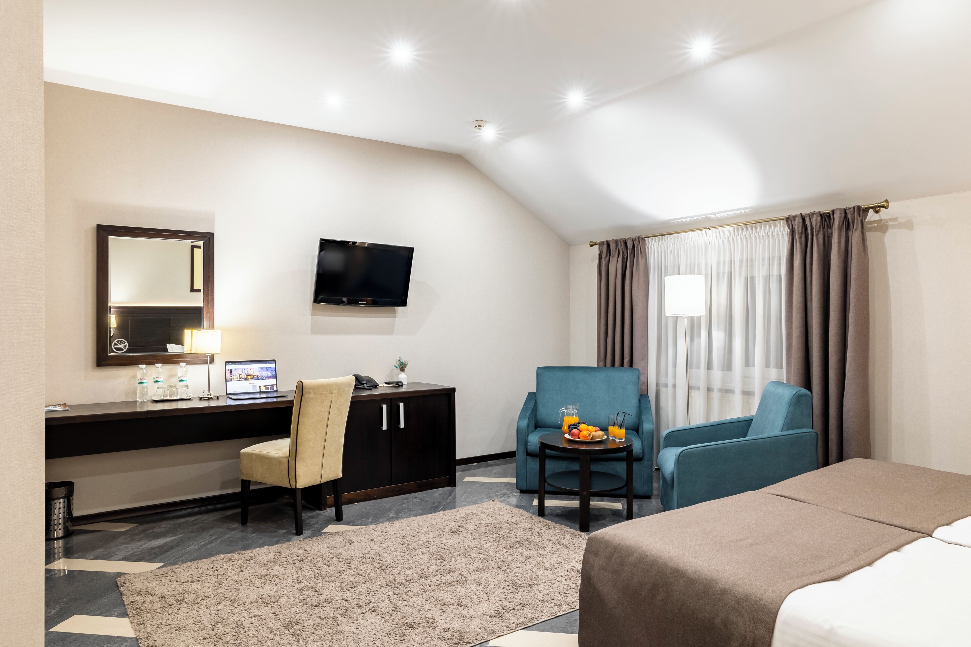 Habitación de hotel con escritorio, dos sillas, cama, televisor y ventana con cortinas.