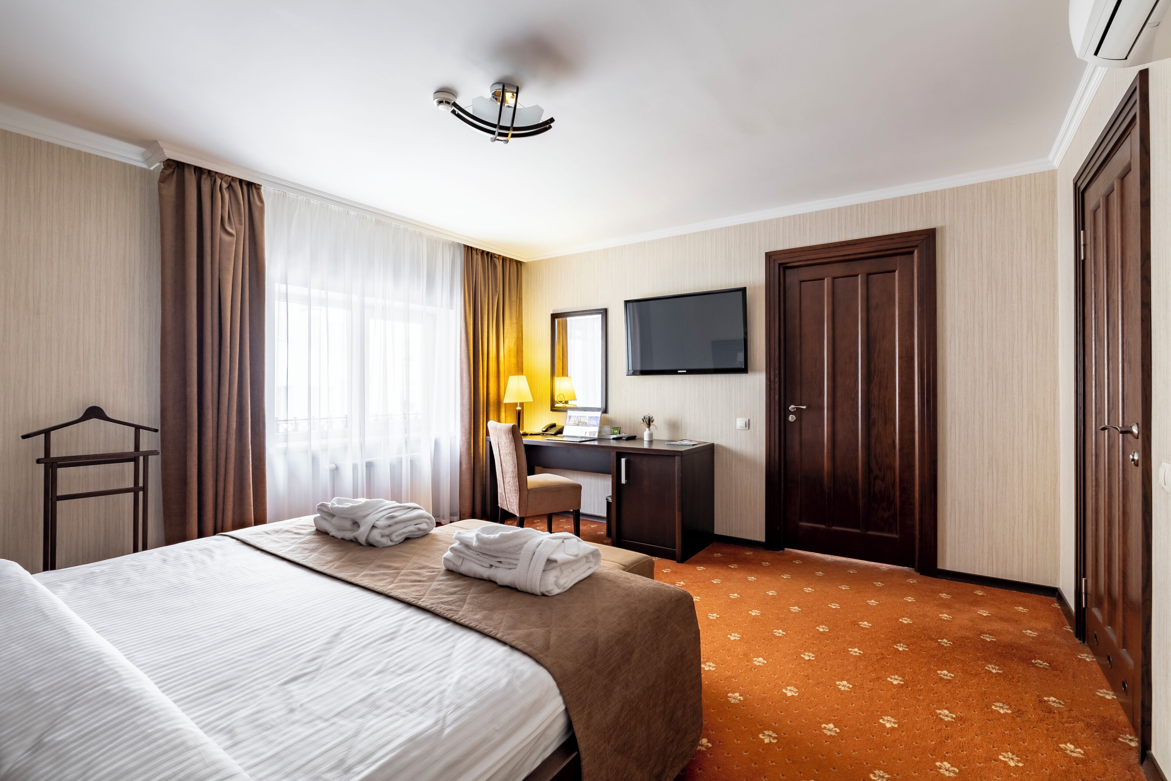 A hotel room with a bed, desk, chair, TV, wardrobe, and a patterned carpet.