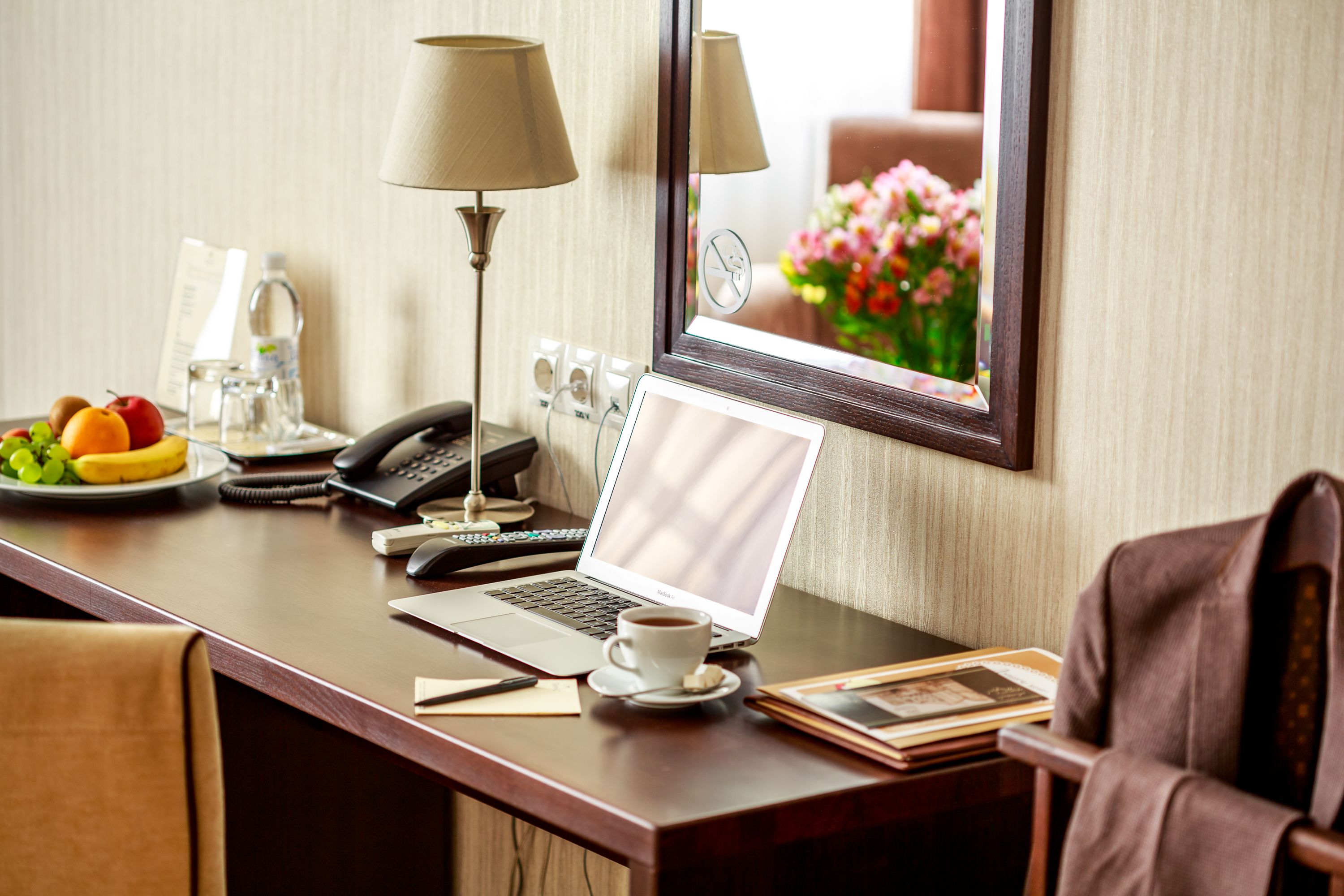 A hotel room desk with a laptop, a cup of coffee, a fruit bowl, a lamp, a phone, and a mirror reflecting a flower arrangement.