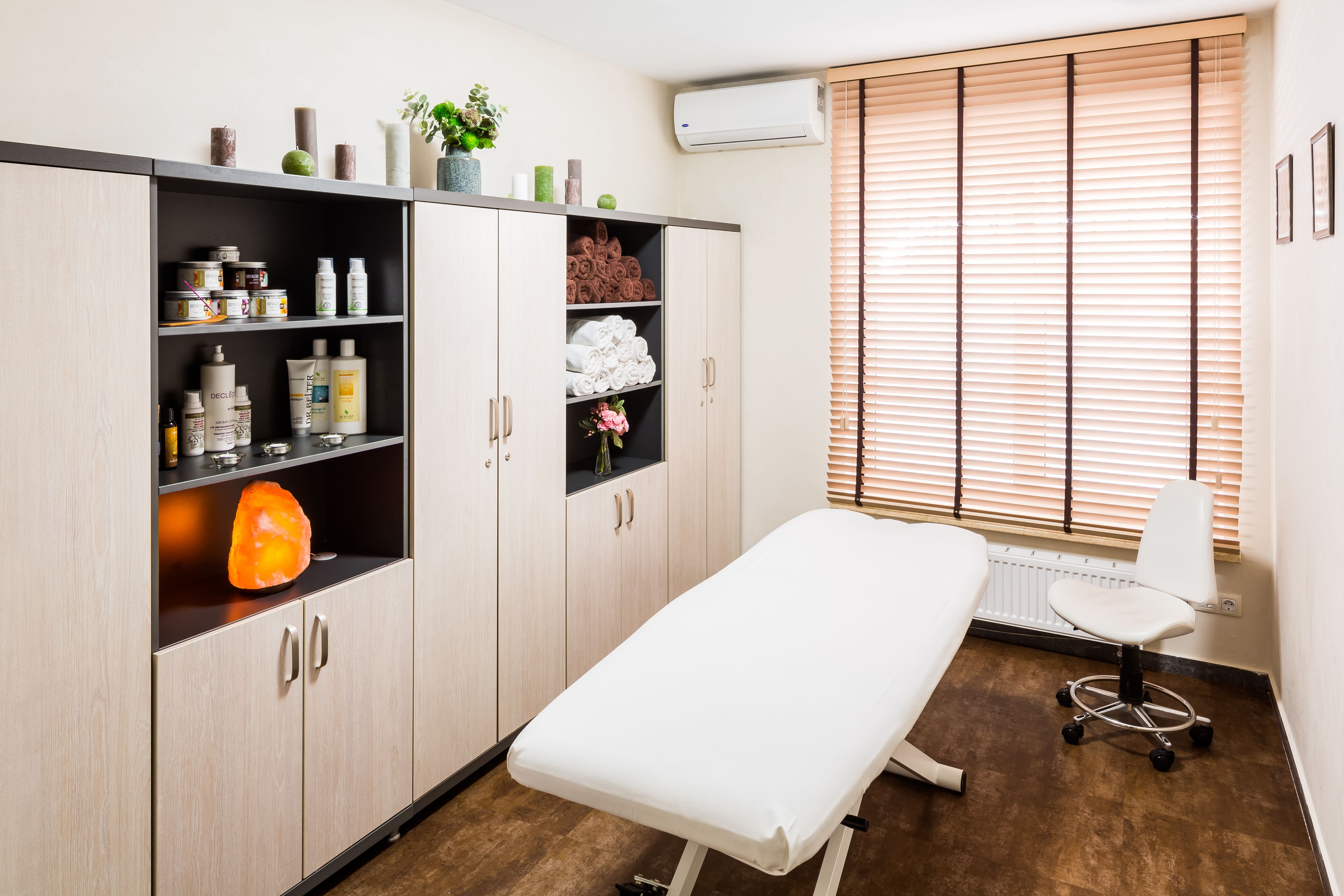 A clinical examination room with a white examination table, a white chair, and shelves with medical supplies.