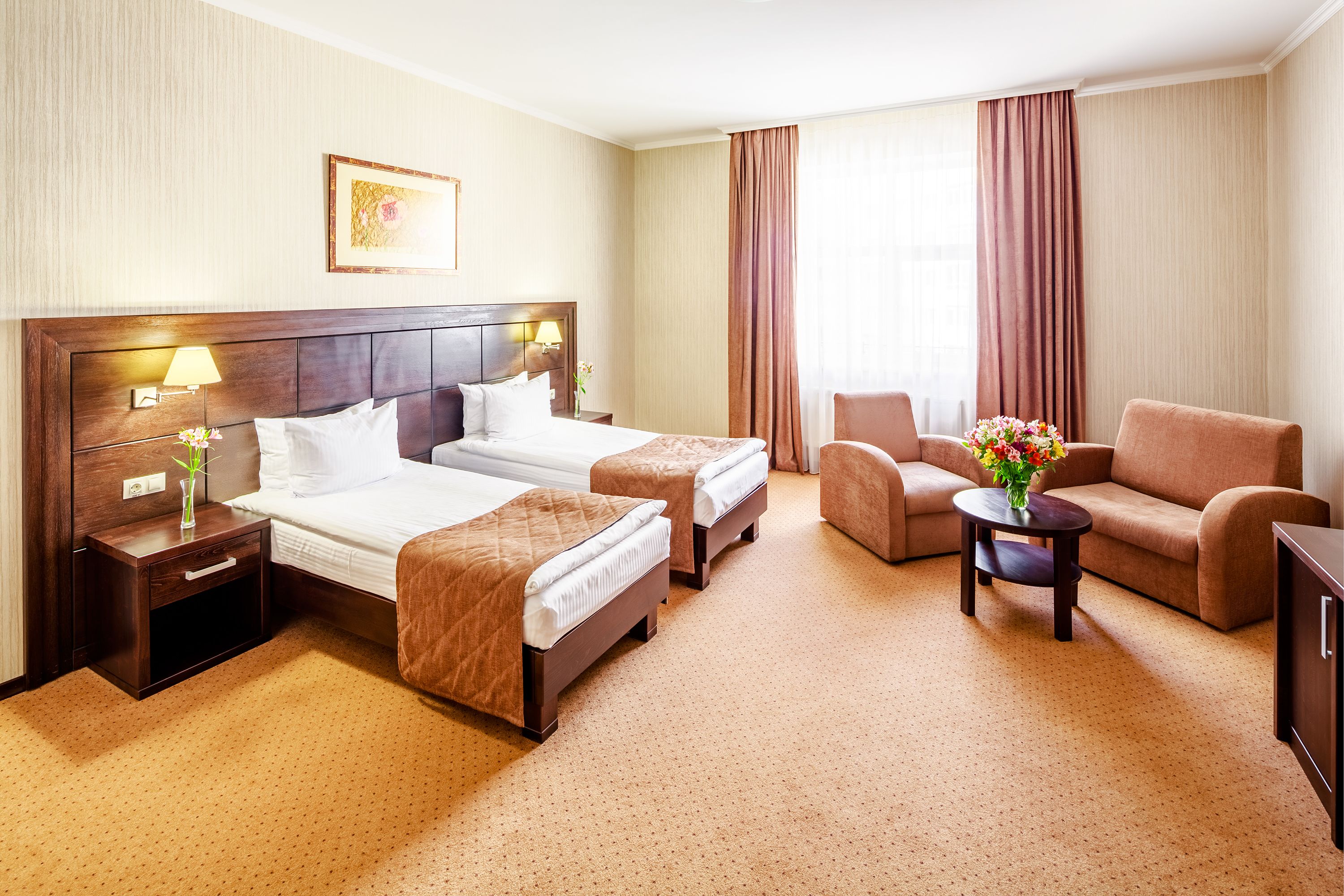 Quarto de hotel com duas camas, um sofá, uma mesinha com flores e uma mesa de cabeceira com abajur.
