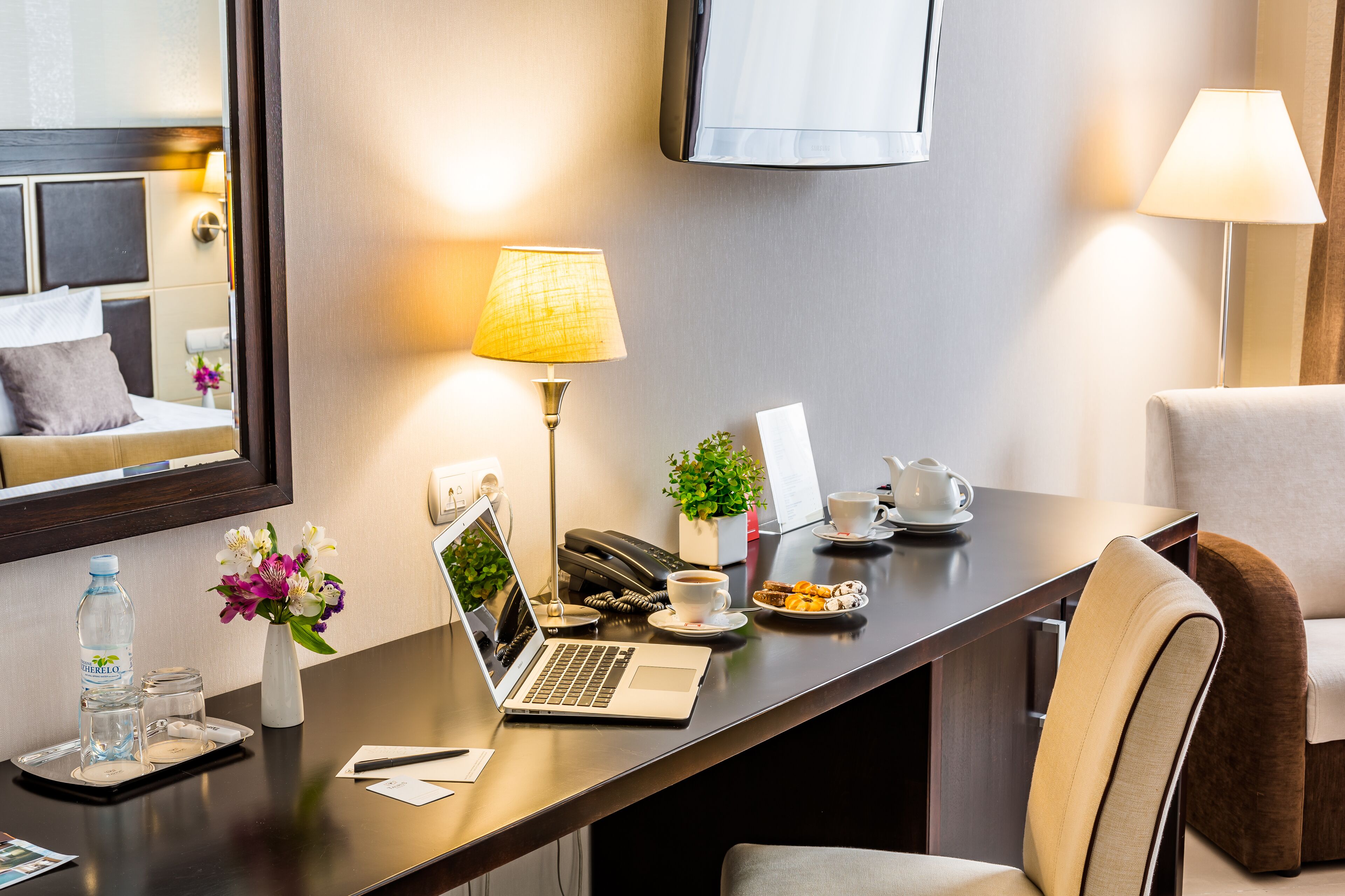 A hotel room with a desk, laptop, coffee cup, and a bed with pillows.