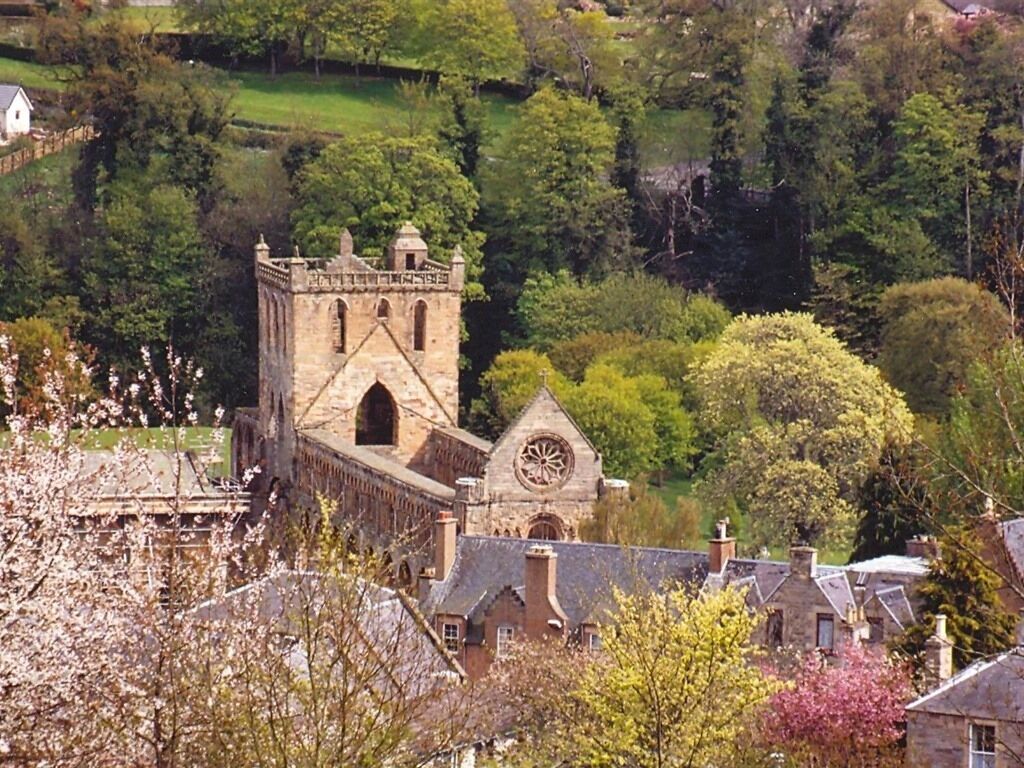 A historic stone building with a clock tower, surrounded by lush greenery and residential houses.