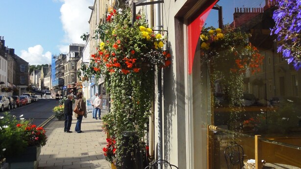 Une vue de rue avec un café doté d’une terrasse et de jardinières suspendues remplies de fleurs.