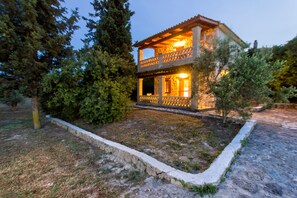 Une maison en pierre à deux étages, dotée d’un balcon, entourée d’arbres et bordée d’un chemin en pierre.