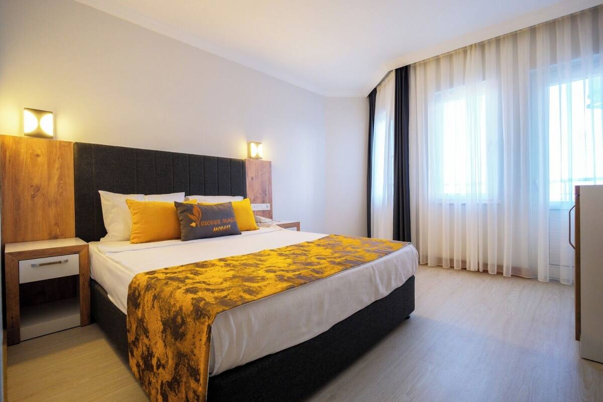 A hotel room with a large bed, two bedside tables, a wooden headboard, and a view of the outside through sheer curtains.