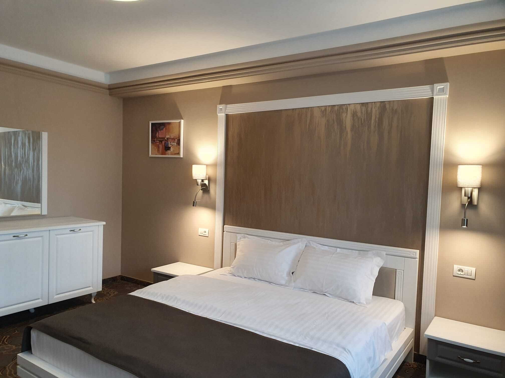 A hotel room with a large bed, a bedside table, a nightstand, a dresser, and two wall-mounted lamps.