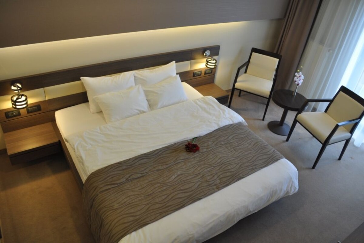 Une chambre d’hôtel avec un grand lit, deux tables de chevet avec des lampes, une chaise et une petite table sur laquelle est posé un vase.