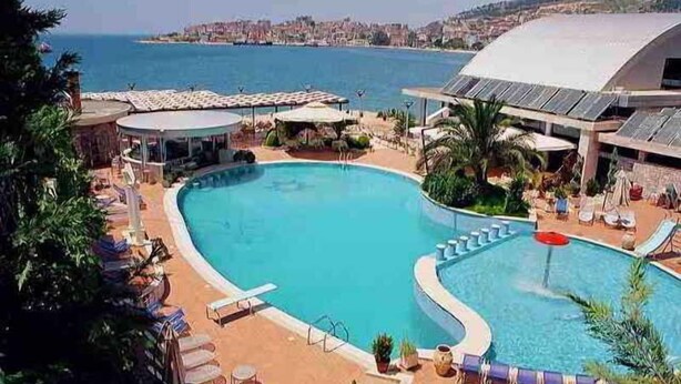 Una piscina de borde curvo, rodeada de sillones de descanso y sombrillas, con vistas al mar y un paisaje urbano de fondo.