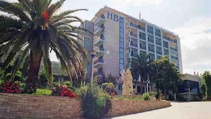 Un hotel con fachada moderna, un acceso ajardinado con palmeras y flores, y un cielo despejado.