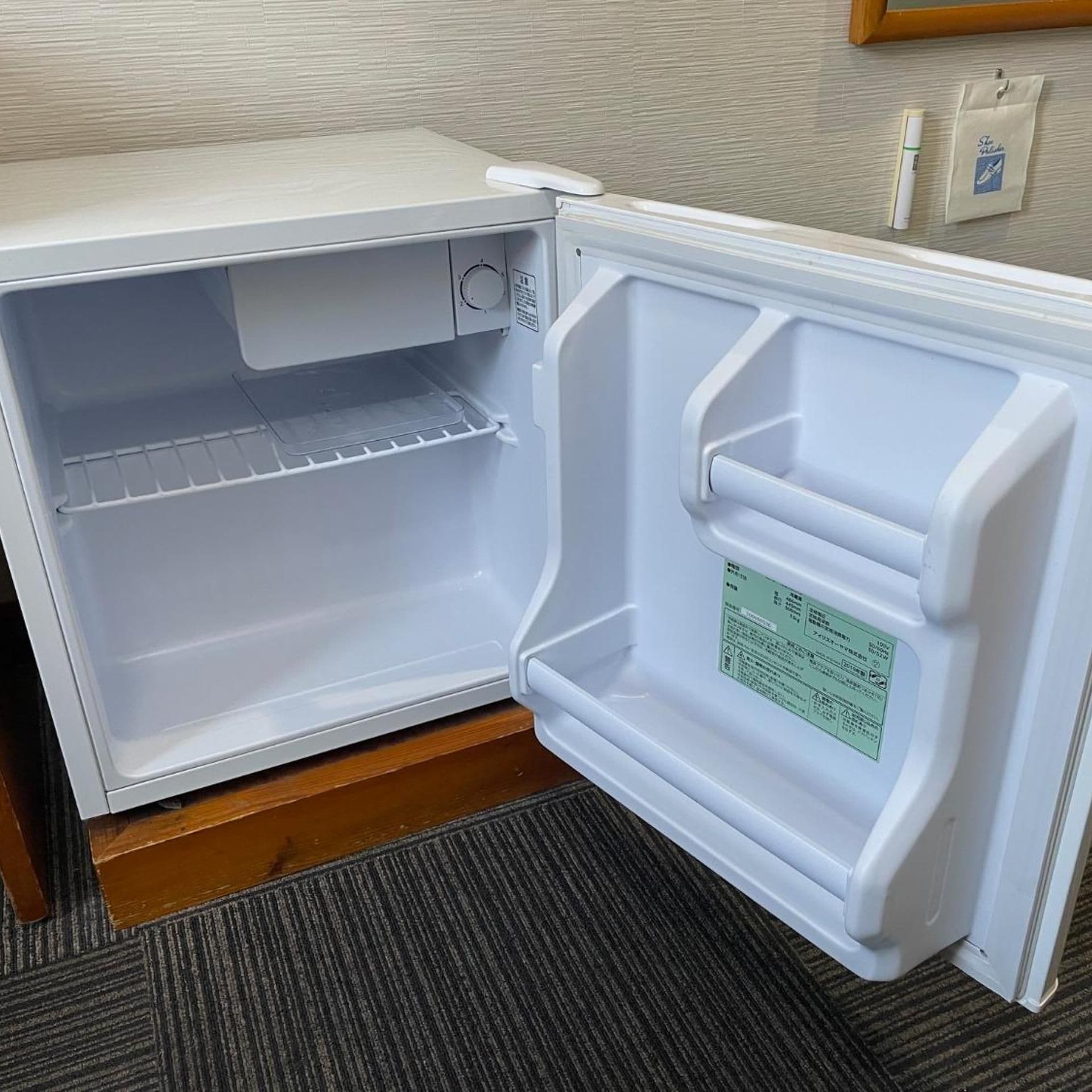 Un refrigerador abierto con un panel de control a la izquierda, una etiqueta verde en la puerta derecha y un piso de madera debajo.