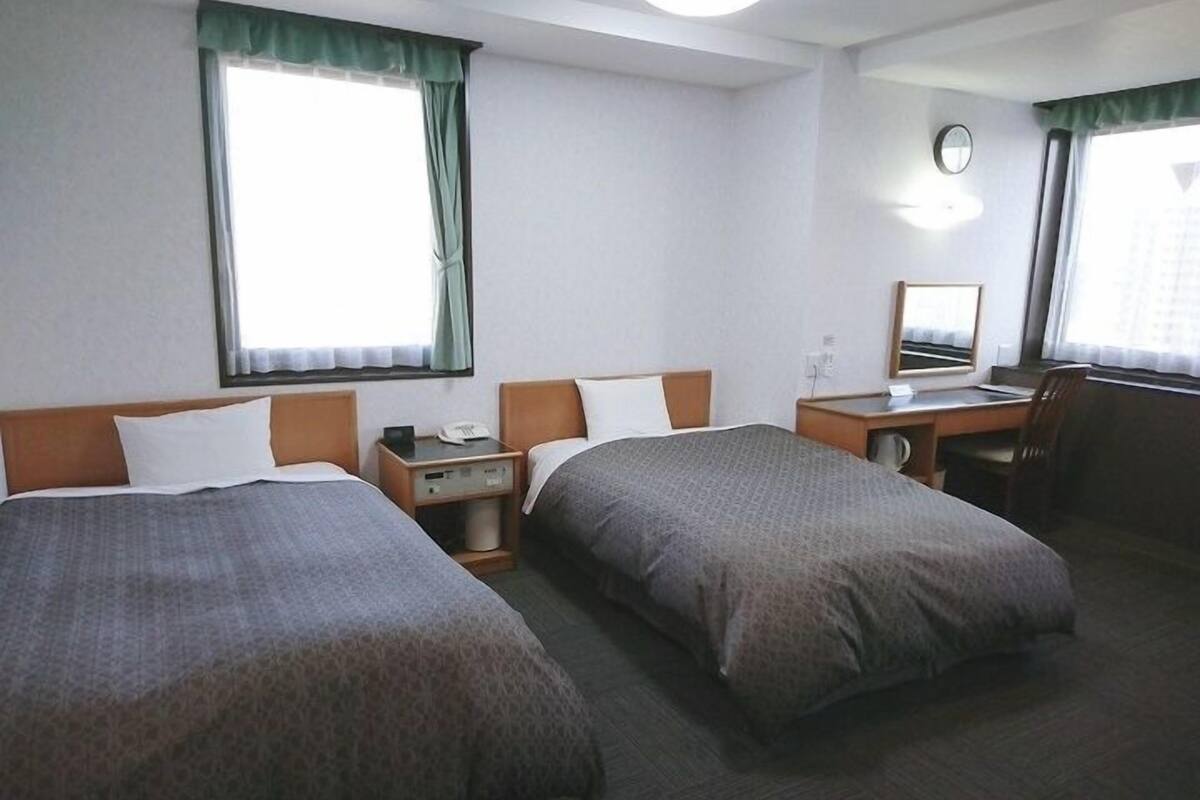 A hotel room with two beds, a desk, a chair, a mirror, and a window with curtains.