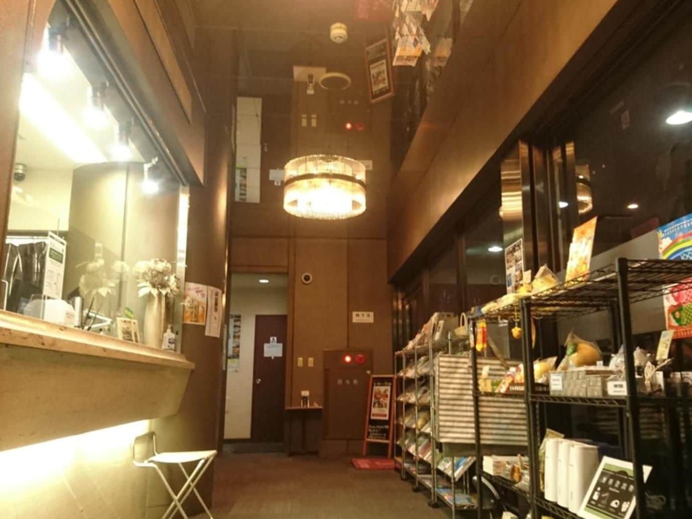 A narrow corridor with a reception desk on the left, a chandelier hanging from the ceiling, and shelves stocked with various items on the right.