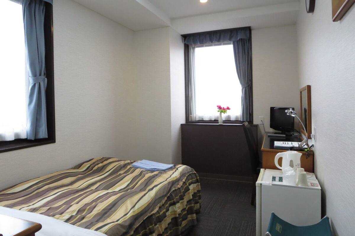 A hotel room with a bed, a desk with a computer, a small kitchenette, and a window with curtains.