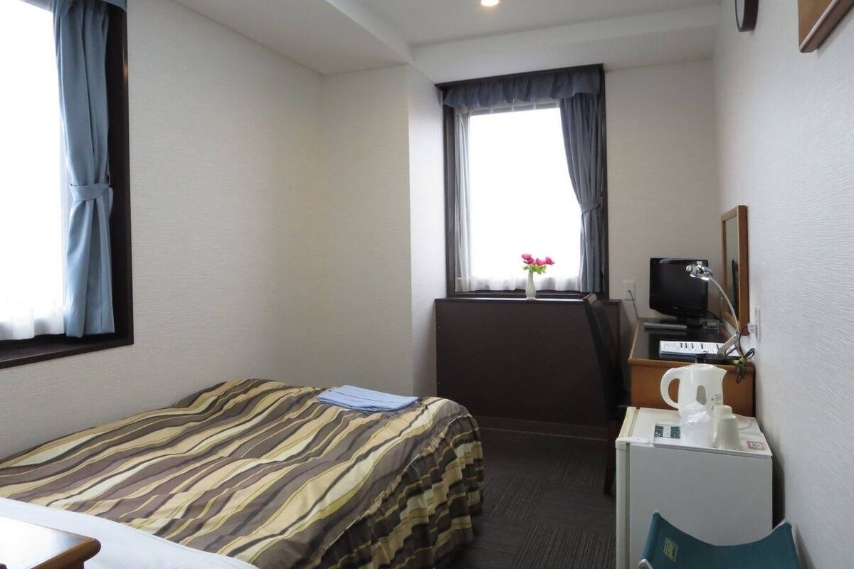 A hotel room with a bed, a desk with a computer, a coffee maker, and a window with curtains.