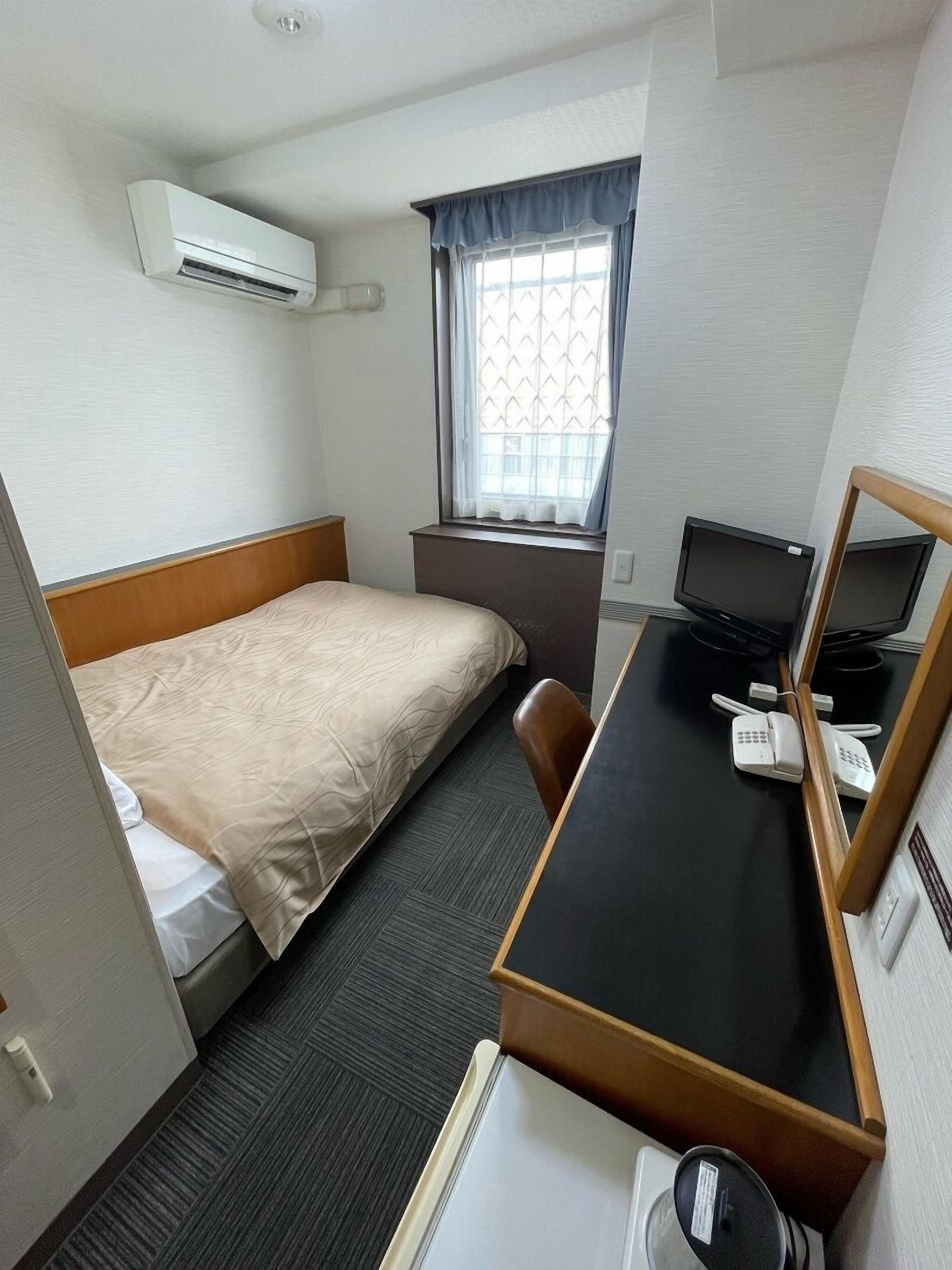 Ein kleines Hotelzimmer mit einem Einzelbett, einem Schreibtisch mit Computer und Telefon, einem Fenster mit Vorhängen sowie einer Klimaanlage.