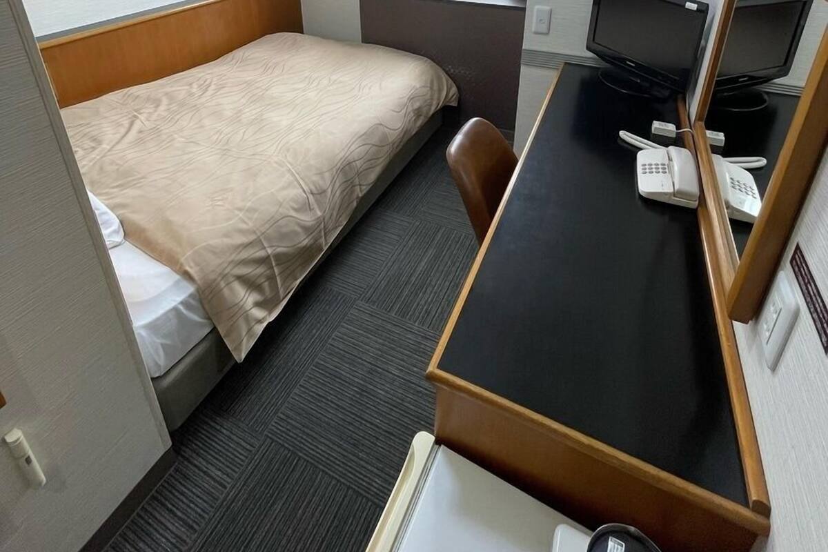A compact hotel room with a single bed, a desk with a computer and phone, a window with curtains, and an air conditioning unit.