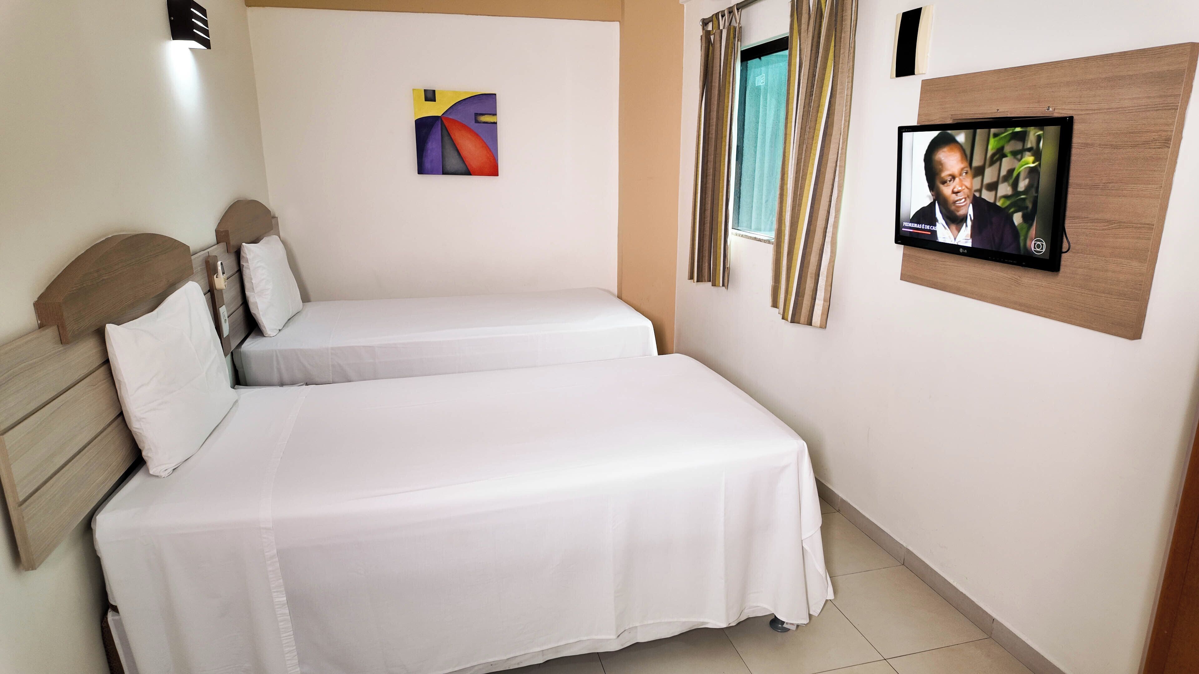 A hotel room with two single beds, a TV on the wall, a painting, and a window with curtains.