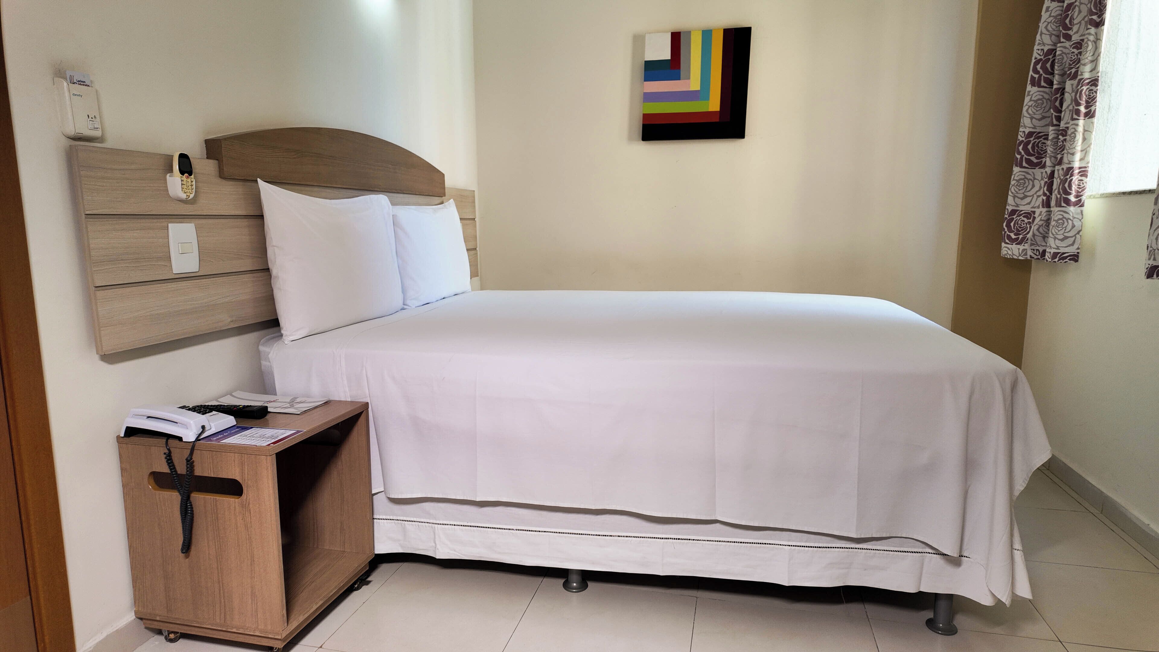 Un lit bien fait dans une chambre d’hôtel, avec une tête de lit en bois, une petite table de chevet en bois sur laquelle se trouve un téléphone, et une peinture abstraite aux couleurs vives accrochée au mur.