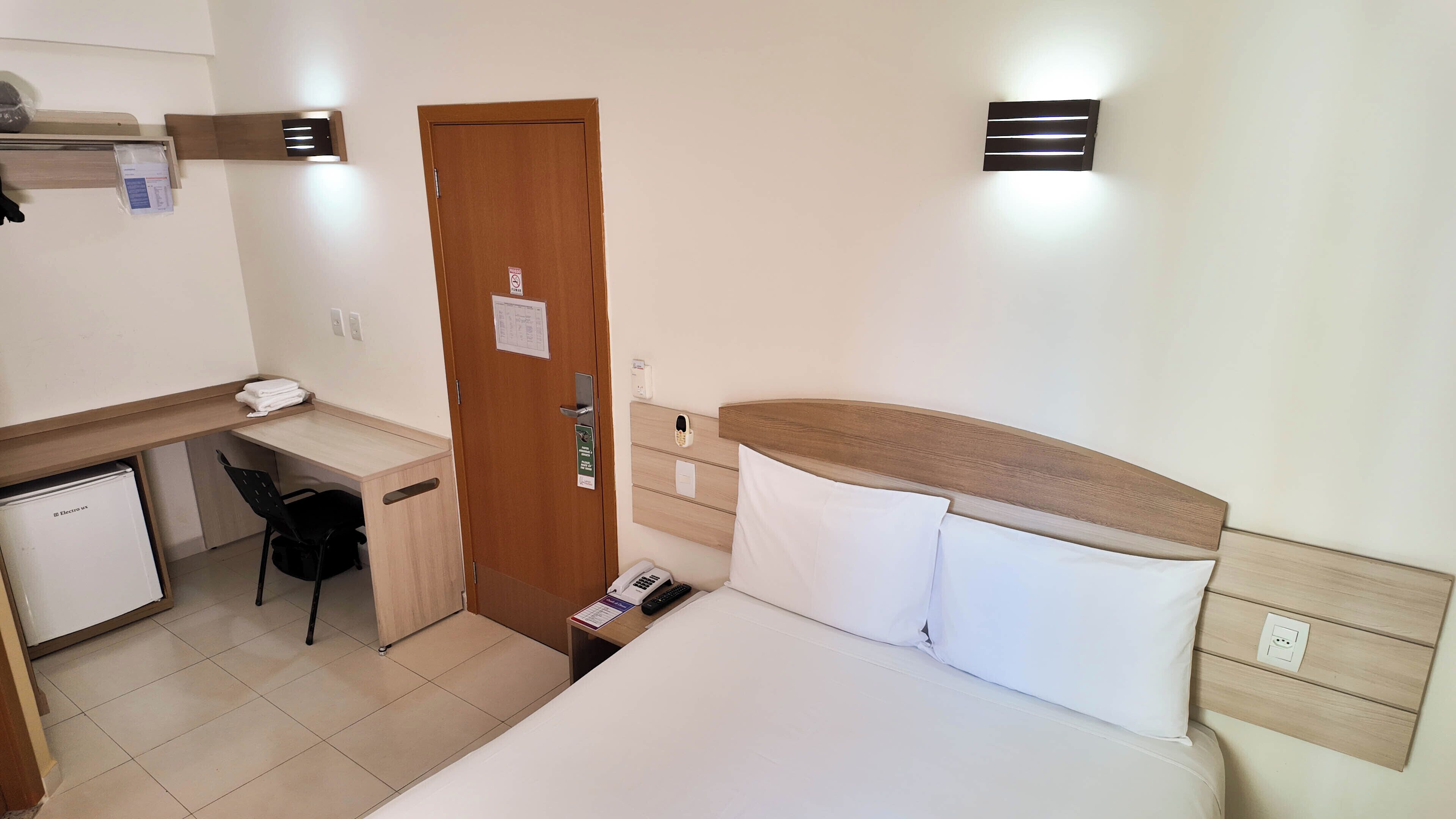 A hotel room with a bed, a desk, a chair, a small refrigerator, and a door with a lock.