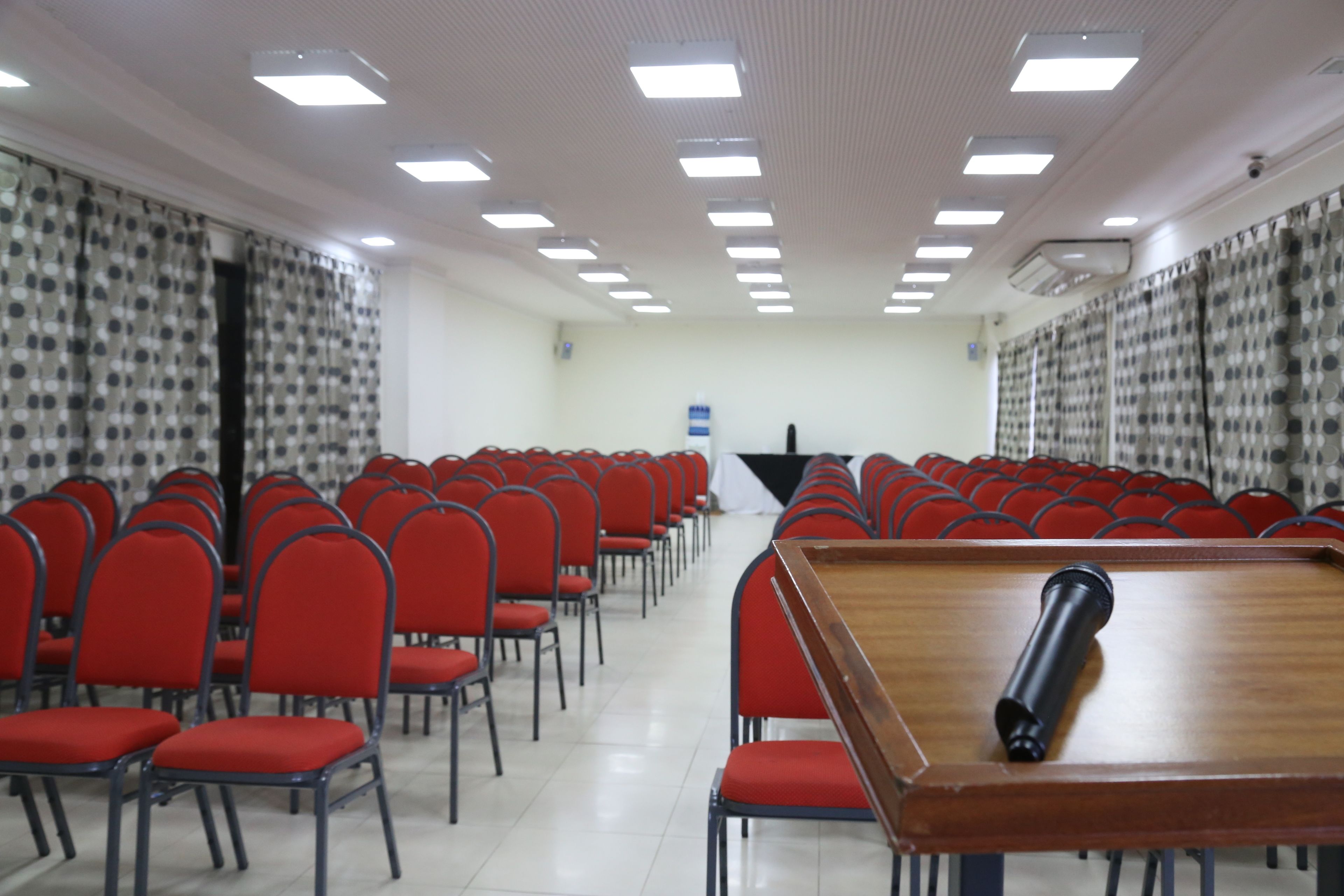 A conference room with rows of red chairs, a wooden table, and ceiling lights.