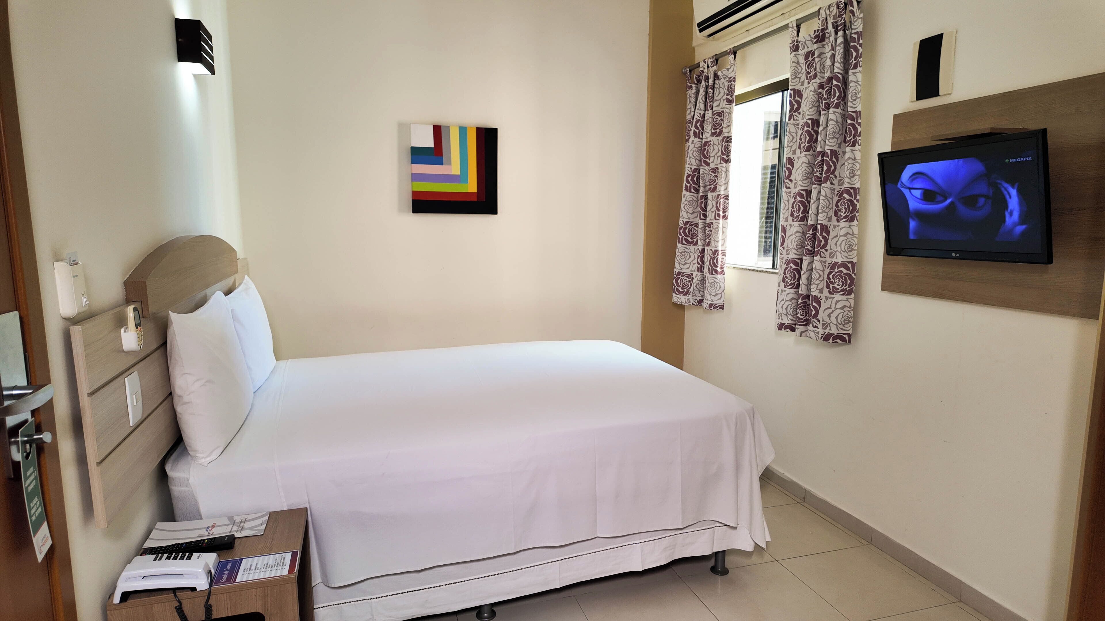 Habitación de hotel con una cama, un televisor montado en la pared, un cuadro y una ventana con cortinas.