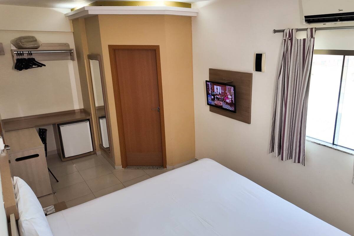 Una habitación de hotel con cama, televisor montado en la pared, ventana con cortinas, puerta y un pequeño escritorio con silla.