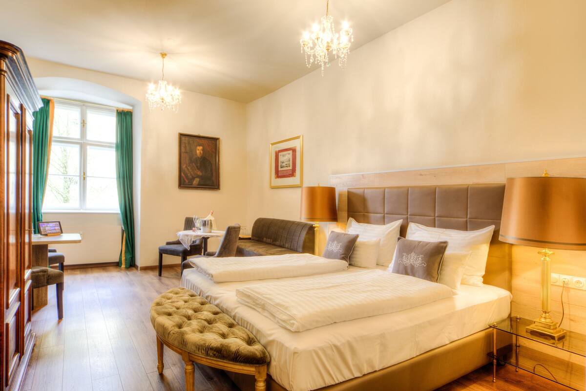 A hotel room with two beds, a desk, a chair, a chandelier, and a window with curtains.