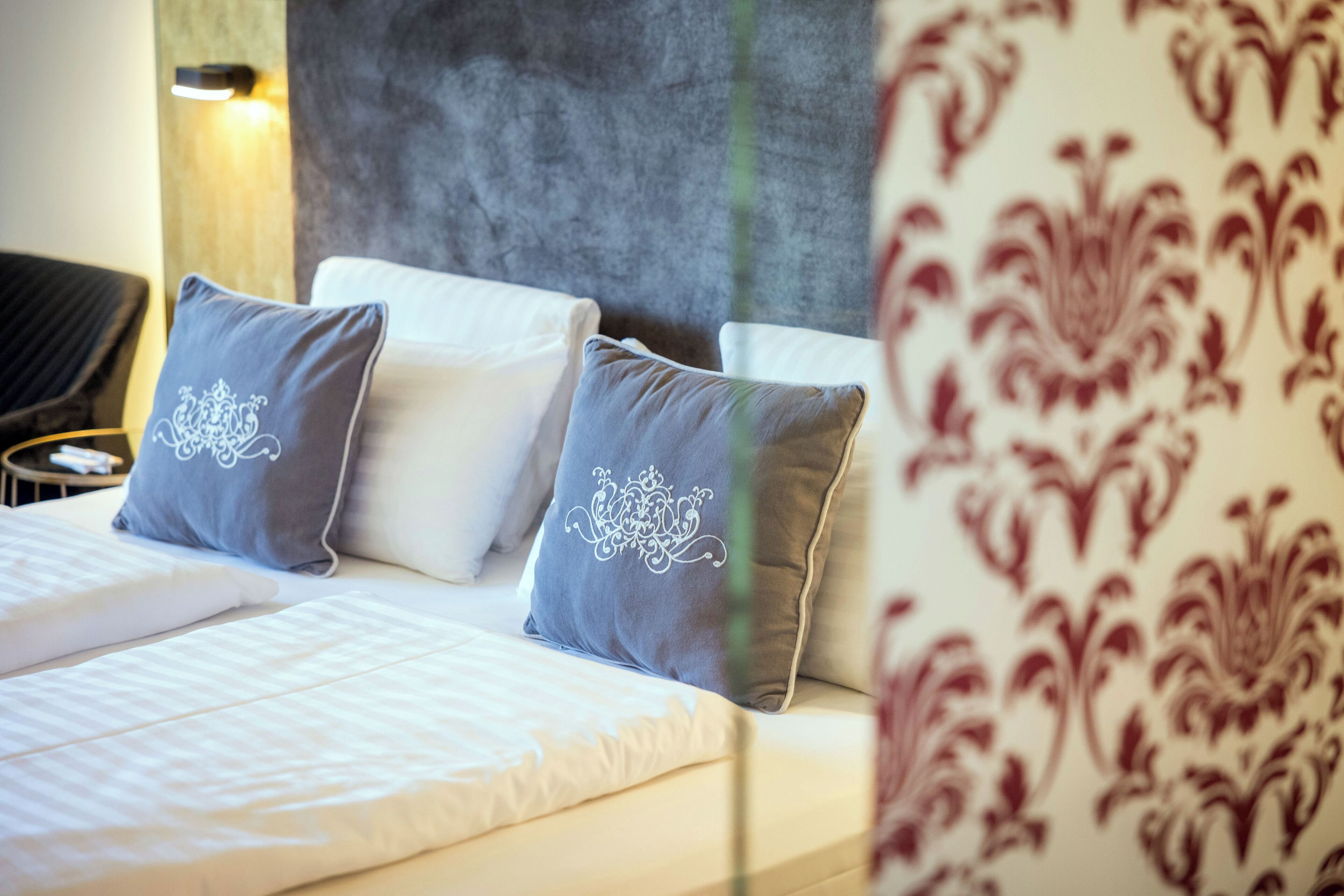 A hotel room with a bed, pillows, a headboard, and a wall with a patterned wallpaper.