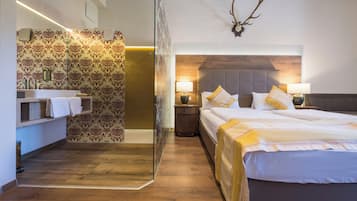 Una habitación de hotel moderna con piso de madera, cabina de ducha de vidrio, cama con ropa blanca y un adorno de cuerno de ciervo en la pared.