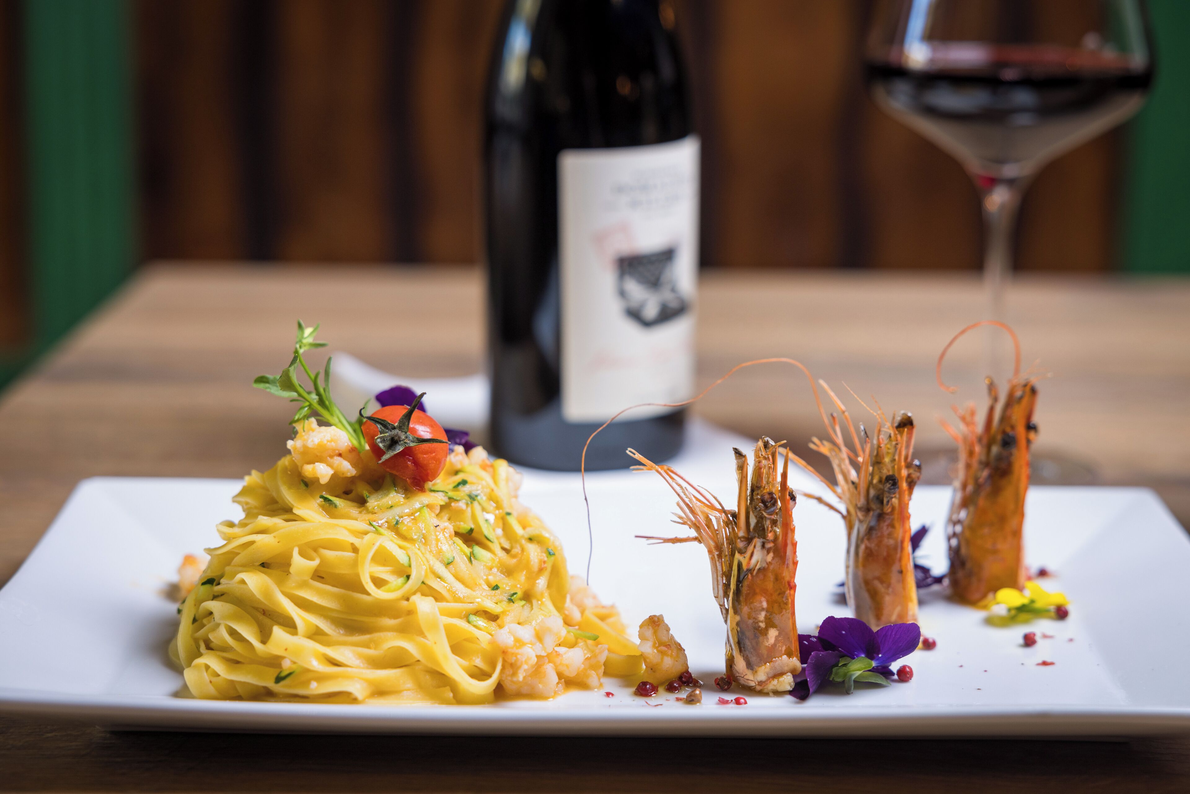 Un piatto di pasta con gamberi e una bottiglia di vino sullo sfondo.
