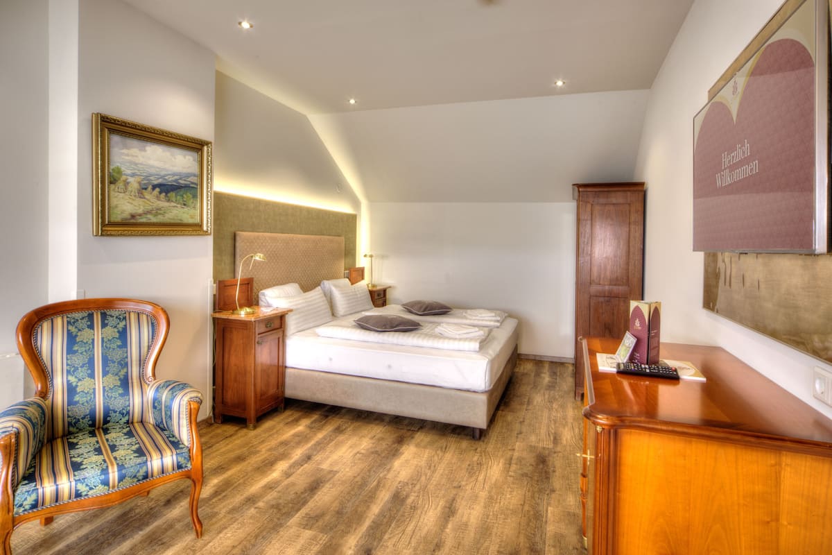 A bedroom with a bed, a nightstand, a chair, a wooden dresser, a painting on the wall, and a welcome sign.
