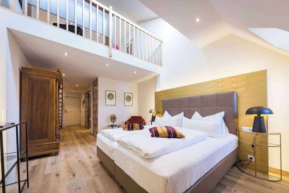A spacious bedroom with a large bed, a wooden wardrobe, a desk with a lamp, and a view of the upper floor.