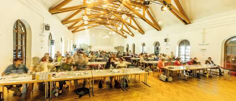 A large hall with high ceilings and wooden beams, furnished with long tables and chairs, suitable for group meetings or dining.