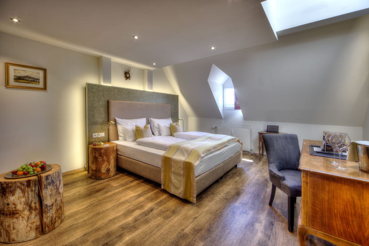 A bedroom with a bed, a desk, a chair, a wooden side table, and a window.