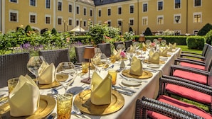Mesa de banquete dispuesta para un evento formal al aire libre, con un edificio amarillo y follaje de fondo.