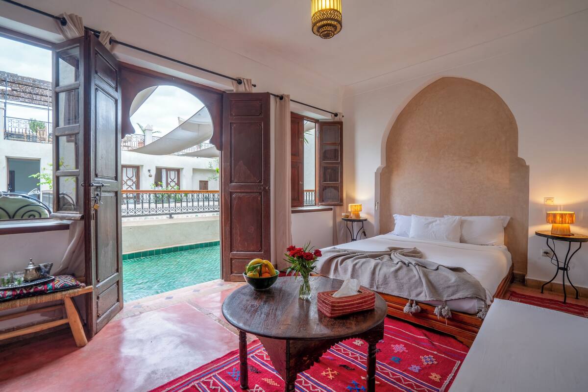 A hotel room with a bed, a small table with a bowl of fruit and flowers, a bedside lamp, and a view of the outside.
