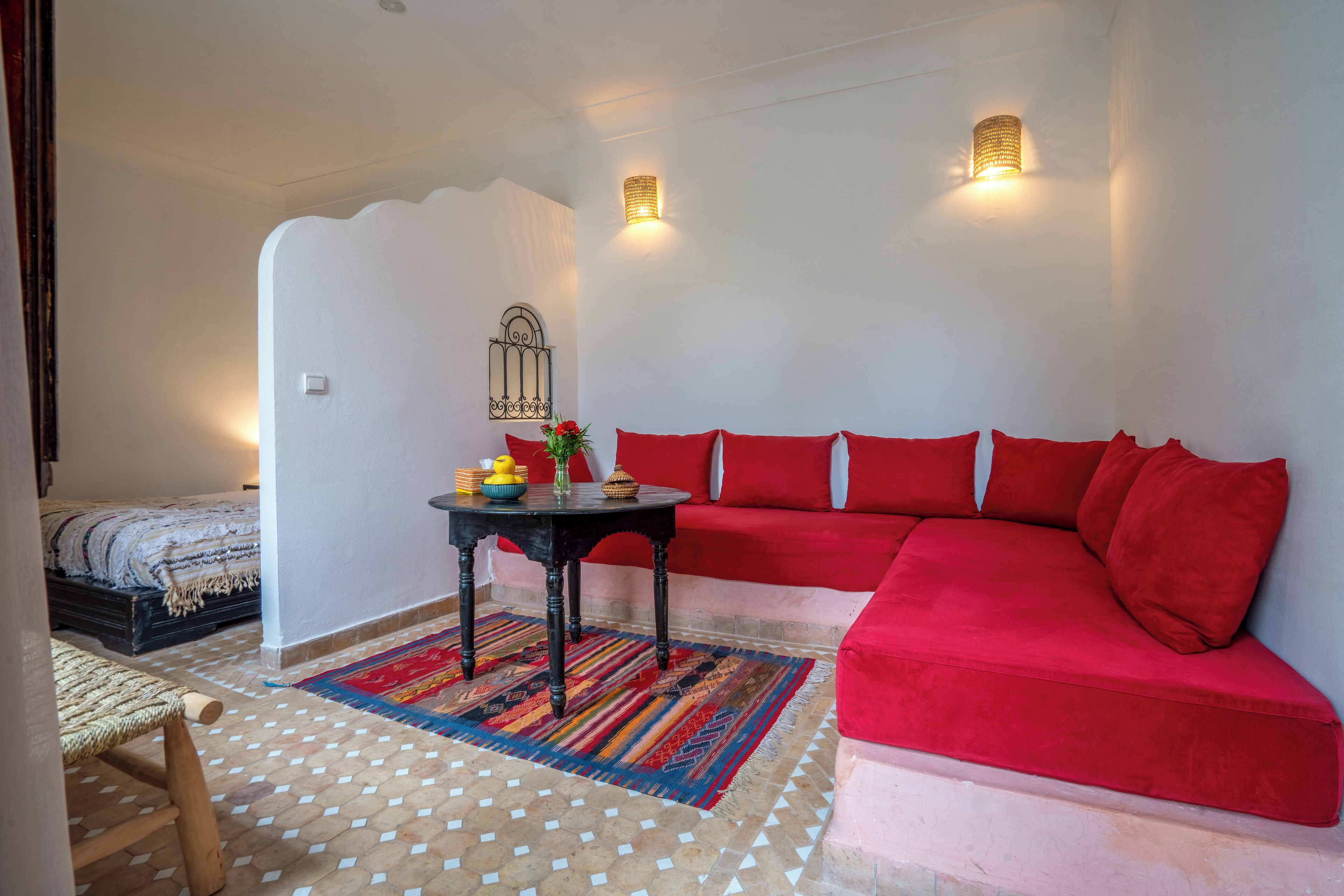Quarto com sofá vermelho, uma mesinha com objetos decorativos, tapete estampado e cama com manta listrada.