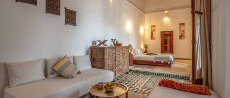 Una habitación amplia con una cama grande, un sofá y una pequeña mesa con fruta. Se observan elementos decorativos en las paredes y una alfombra estampada.