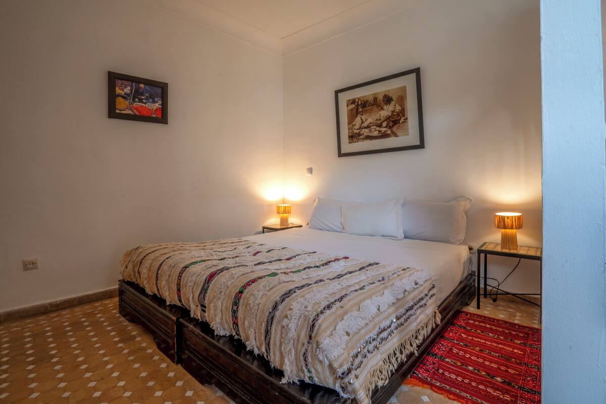 A bedroom with a bed, two bedside lamps, a red rug, and two framed pictures on the wall.