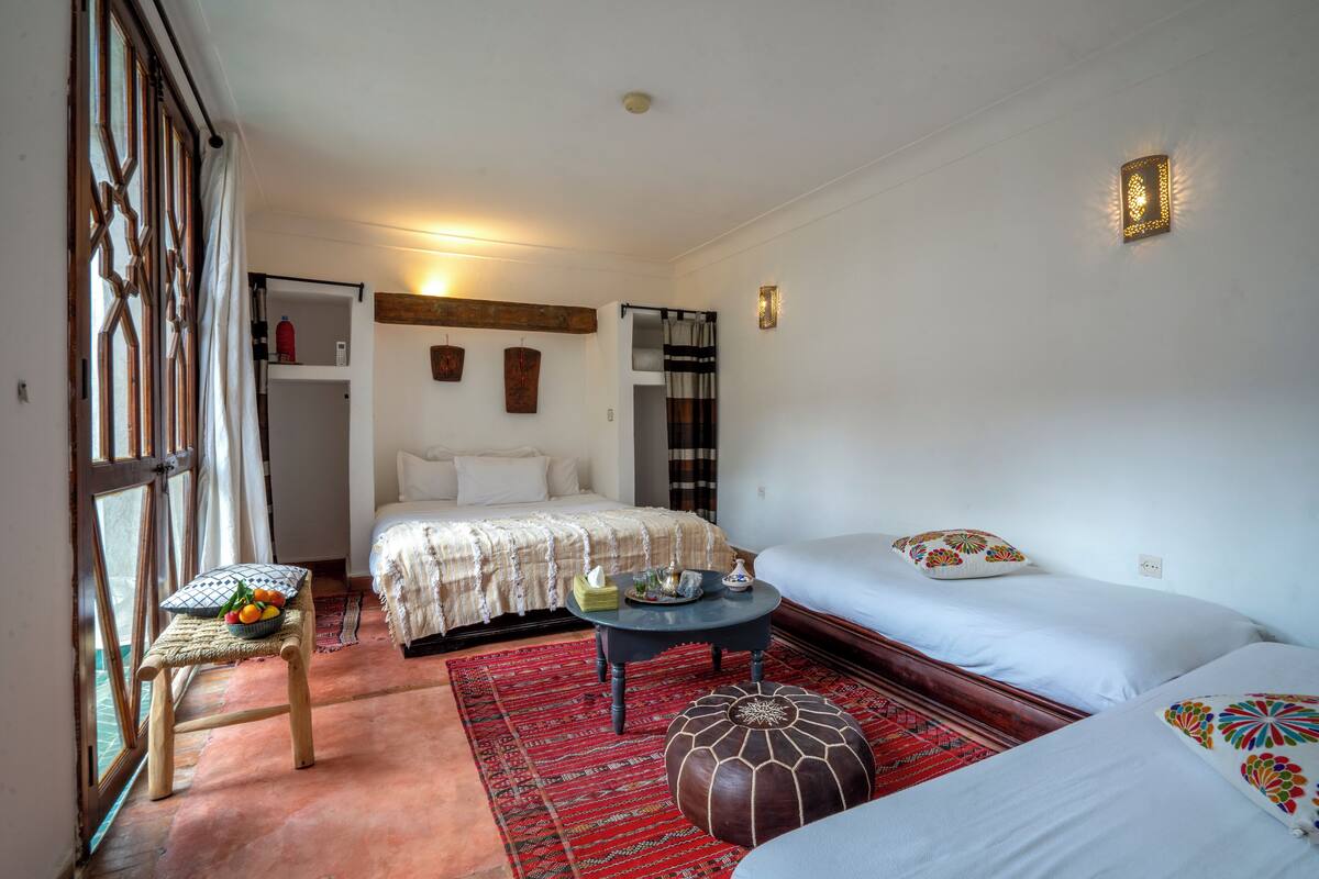 A bedroom with a bed, two bedside tables, a small stool, a red rug, and a window with curtains.