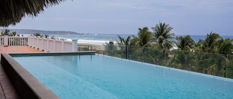 A clear view of a swimming pool with a glass barrier, a beach in the background, and palm trees.