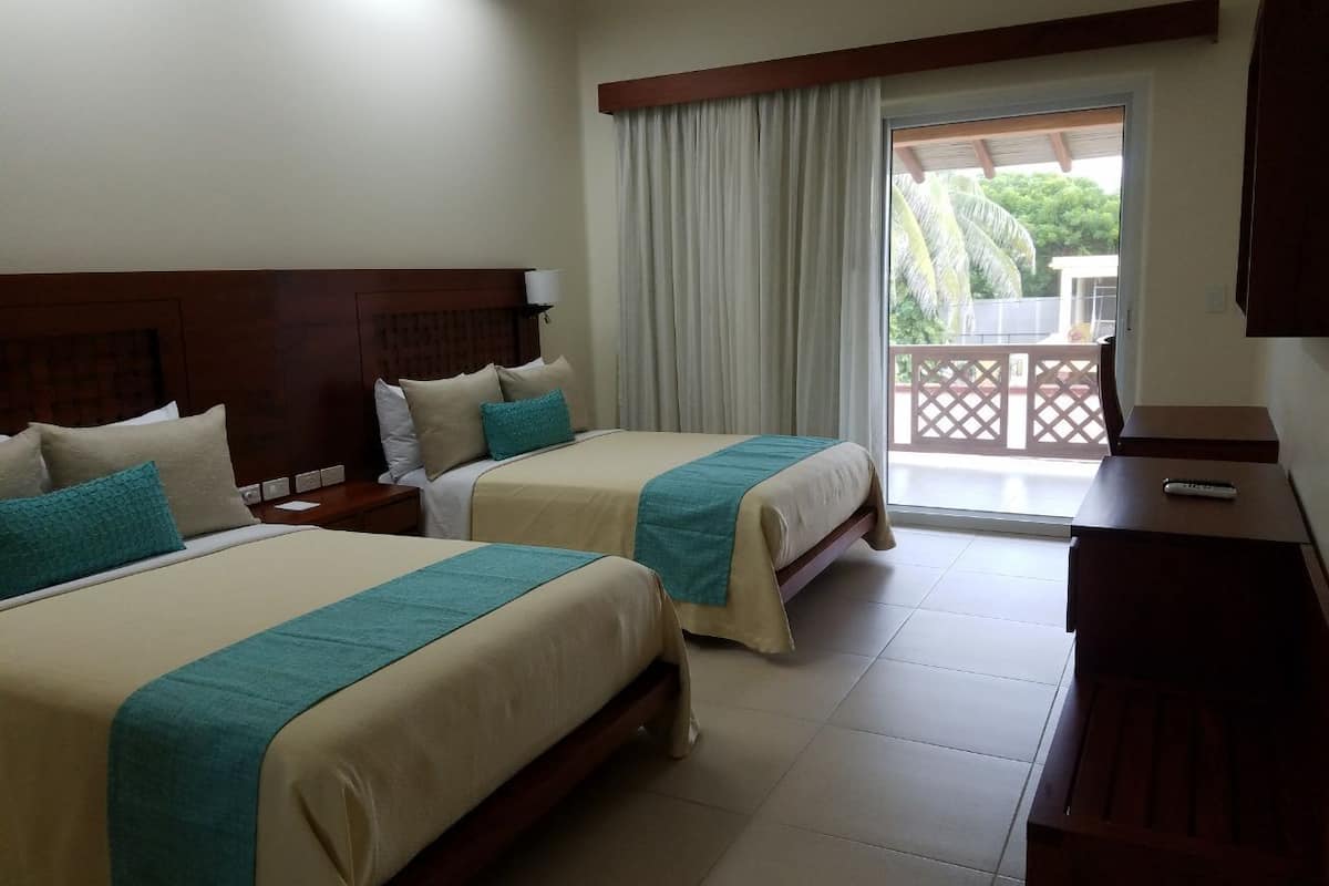 A hotel room with two beds, a desk, a chair, a TV, and a balcony with a view of palm trees.
