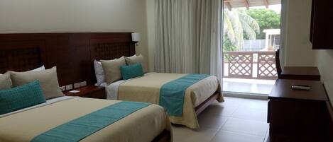 A hotel room with two beds, a desk, a chair, a TV, and a balcony with a view of palm trees.
