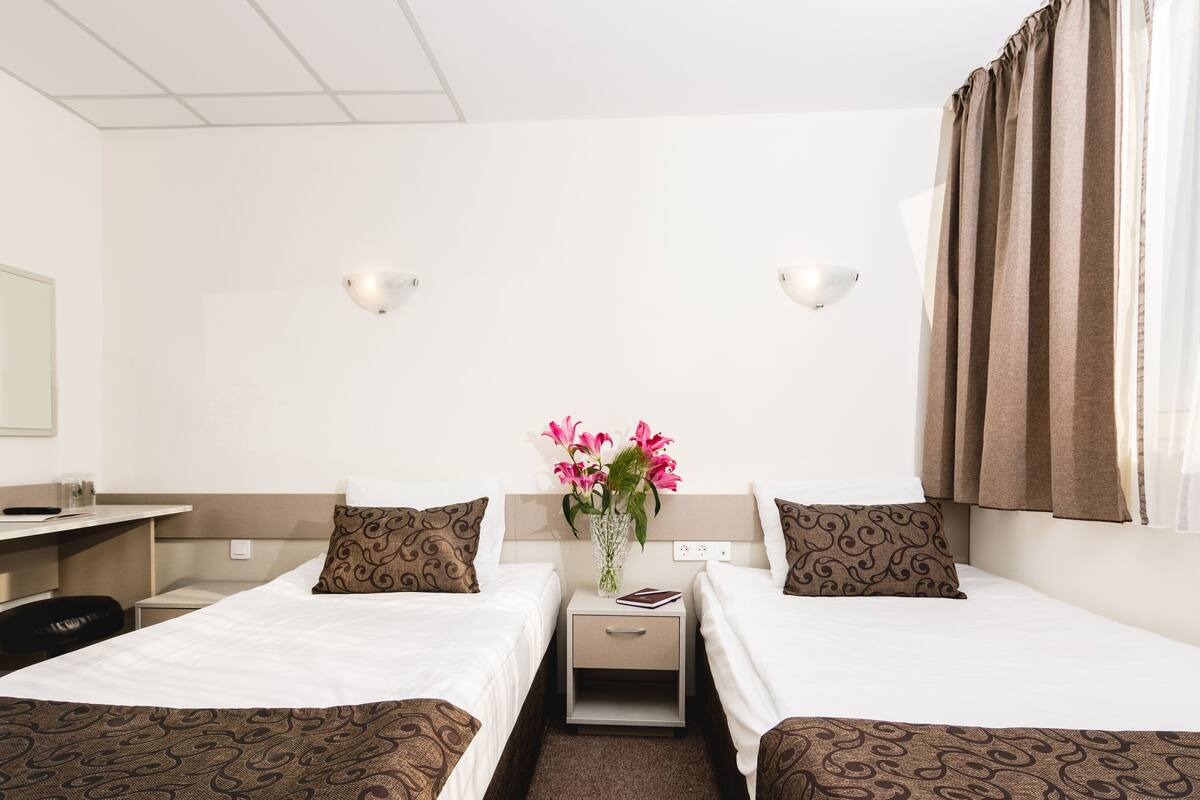 A hotel room with two single beds, a desk, a chair, a nightstand with a vase of flowers, and a window with curtains.