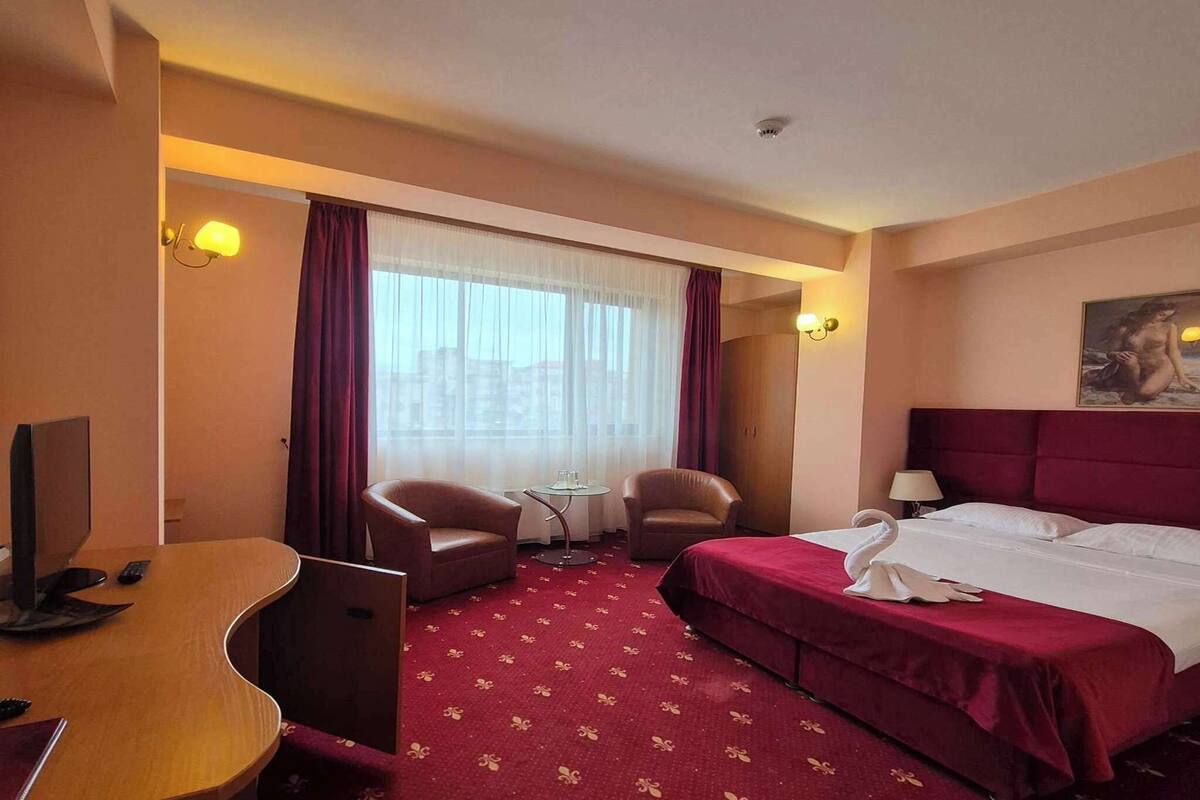 A hotel room with a bed, two armchairs, a desk with a computer, a small table, and a window with curtains.