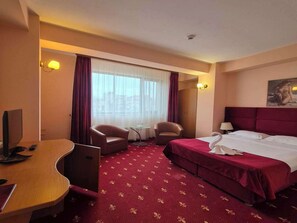 A hotel room with a bed, two armchairs, a desk with a computer, a small table, and a window with curtains.